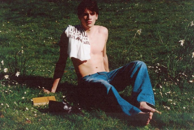 Young adult male poses shirtless in a grassy field, wearing jeans and a white t-shirt draped over his shoulder, bathed in natural light.