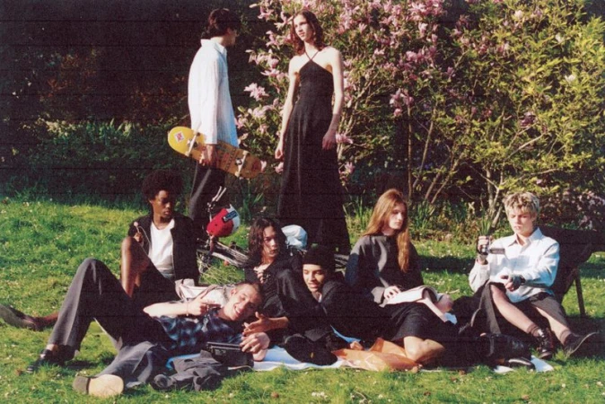 Group of young adults gather outdoors on grass in daytime, with skateboard, bicycle, books, and casual vintage fashion.