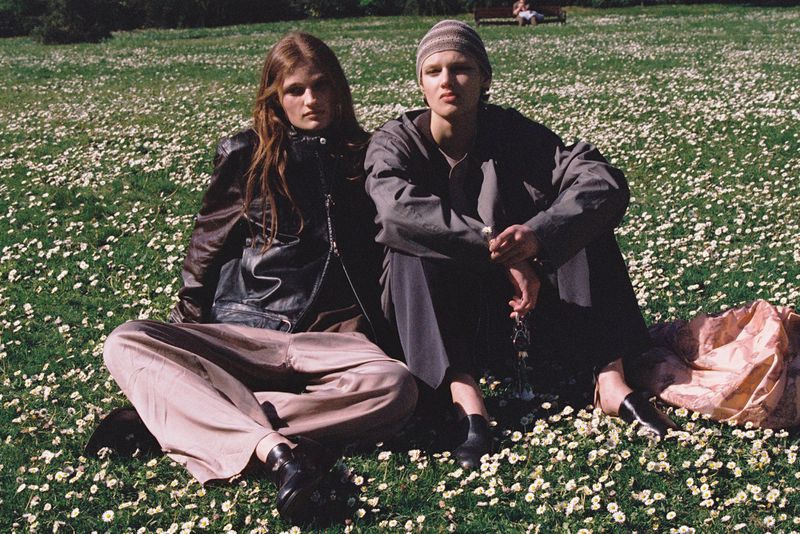 Two people sit on grass covered with daisies. One wears a brown leather jacket, the other a gray shirt and beanie, captured in daylight.