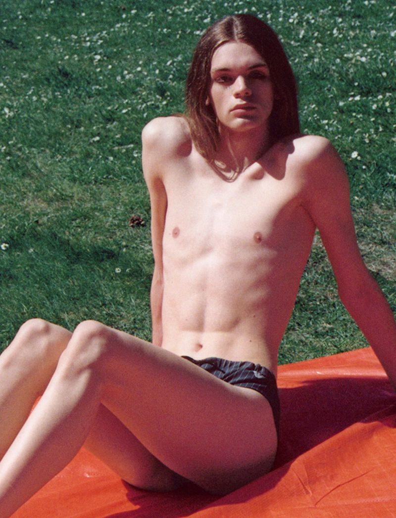 Shirtless young adult male with long hair poses in underwear on an orange sheet outdoors on grass in bright sunlight.