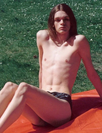 Shirtless young adult male with long hair poses in underwear on an orange sheet outdoors on grass in bright sunlight. 