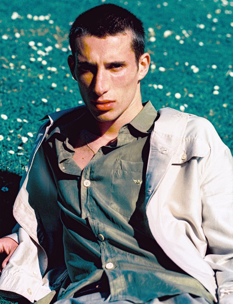 Portrait of a young adult male with short dark hair, wearing a green button-down shirt and a beige jacket, lying in a field of daisies.