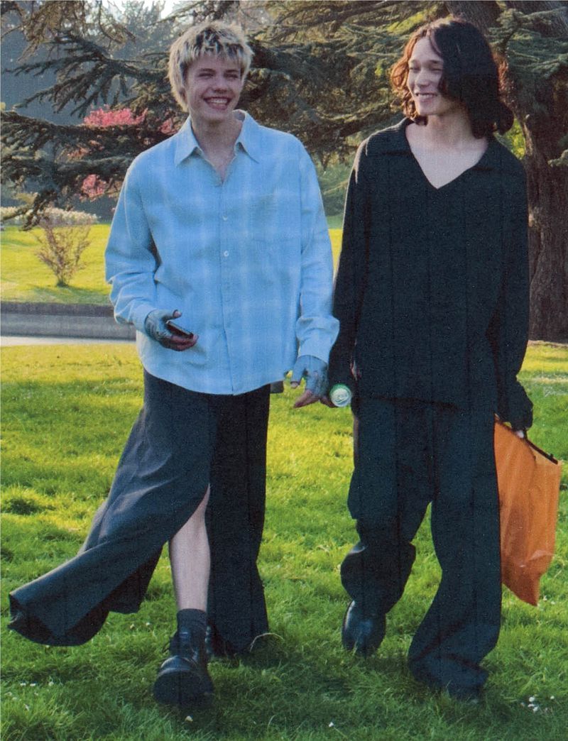 Two young adults stroll on a sunny lawn; one in blue plaid and the other in dark clothing, both smiling and carrying items.