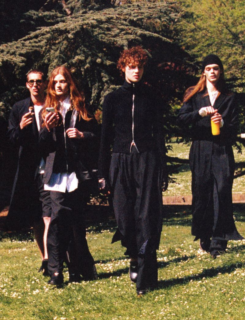 Four people stroll in a park setting, dressed in dark, elegant casual wear, creating a relaxed yet fashionable editorial scene.