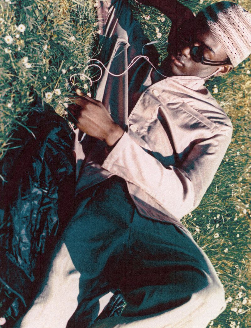 An adult man lies on grass wearing sunglasses and earphones while looking at a cell phone during daytime.