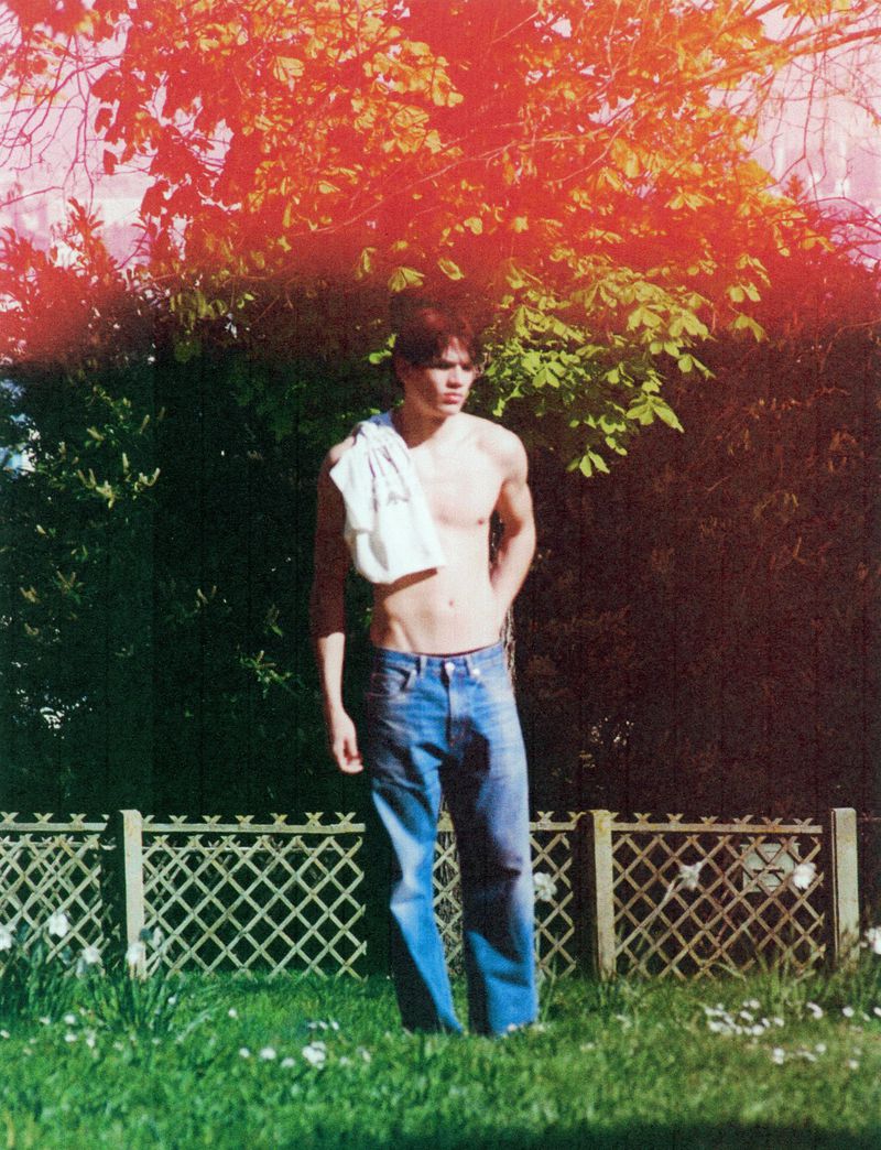 A fair-skinned adult male stands in a grassy yard wearing jeans with a white towel draped over his shoulder, posing in outdoor sunlight.
