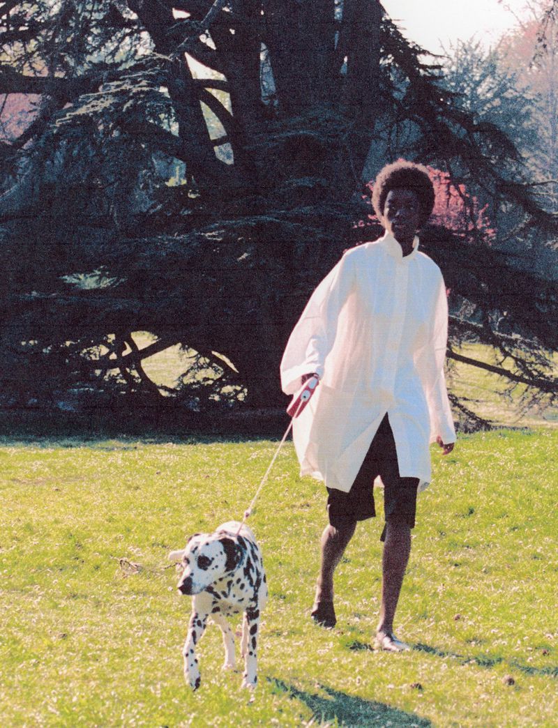 Woman walking a Dalmatian dog in a park. She wears a white shirt and black shorts, enjoying a sunny day in a grassy field.