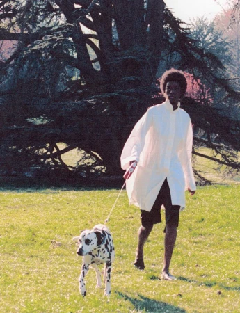 Woman walking a Dalmatian dog in a park. She wears a white shirt and black shorts, enjoying a sunny day in a grassy field.