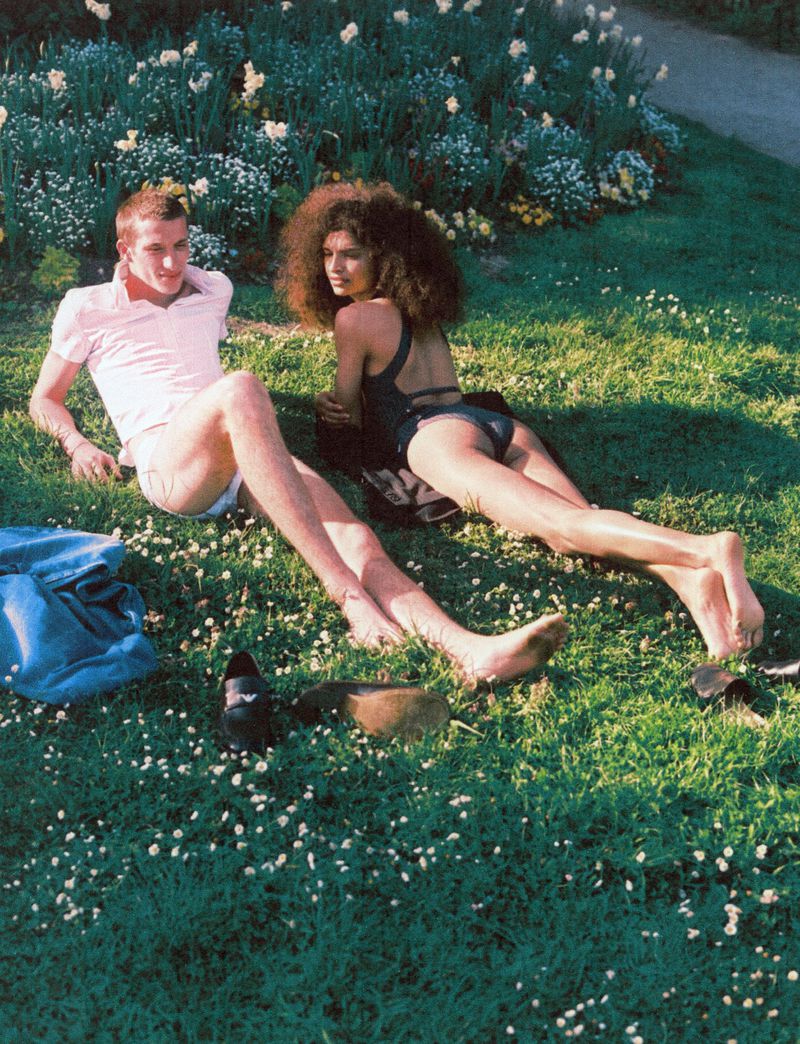 A man and woman, barefoot, casually recline on grass dotted with tiny white flowers, with a floral background and a blue blanket.
