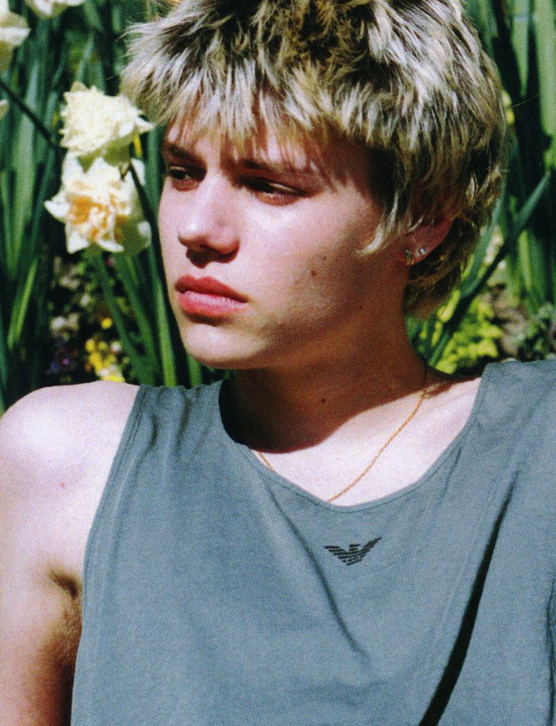 Close-up portrait of a young adult with short blond hair in a sleeveless top against a backdrop of blooming daffodils.