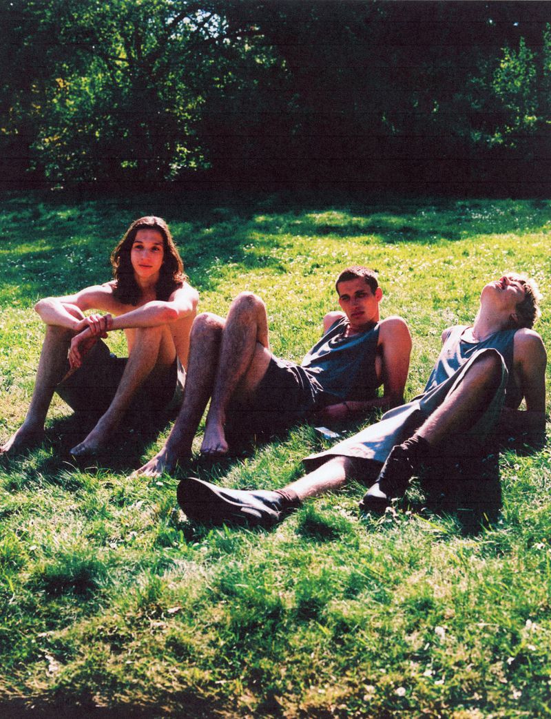 Three young adults are lounging on a grassy lawn, basking in the sunlight filtering through the trees. The outdoor setting evokes a relaxed, summery vibe.
