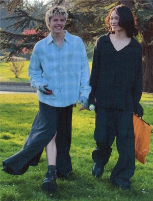 Two young men walking together on a grassy park lawn, one in a light blue patterned shirt and dark long skirt, the other in black.