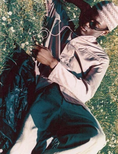 Black man wearing sunglasses and a patterned beanie lying in a grassy field with small white wildflowers