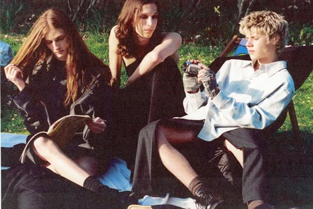 Three models relaxing outdoors on a blanket, with one holding a camcorder and another reading a magazine.
