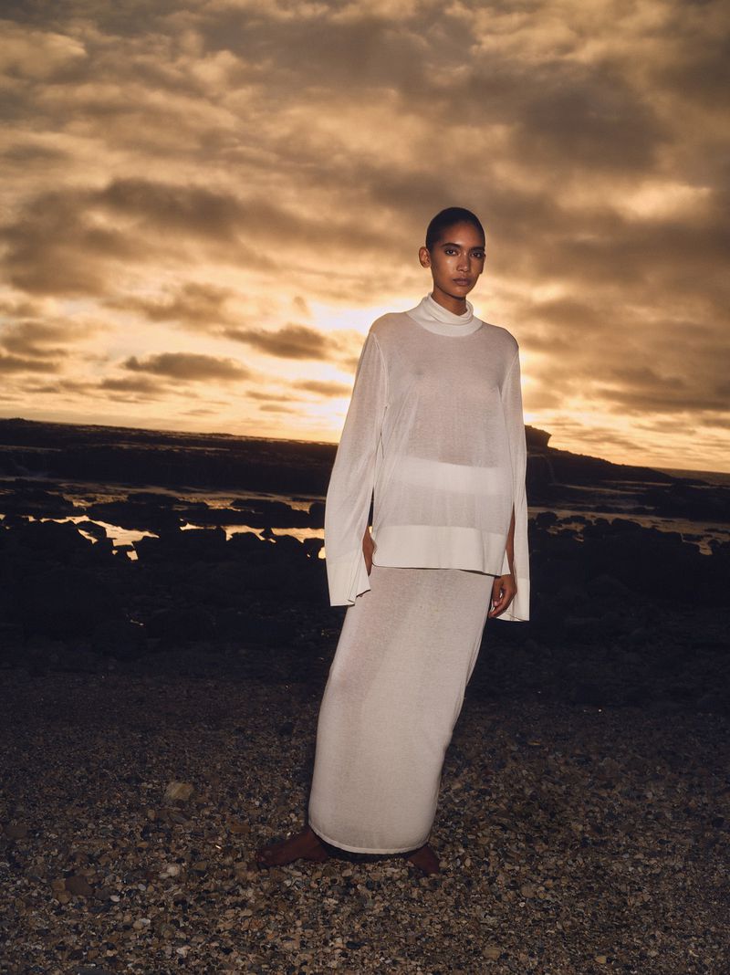 A woman in a white dress stands on a beach at sunset, wearing a sheer, long-sleeved top and a long skirt with a golden cloudy sky behind her.