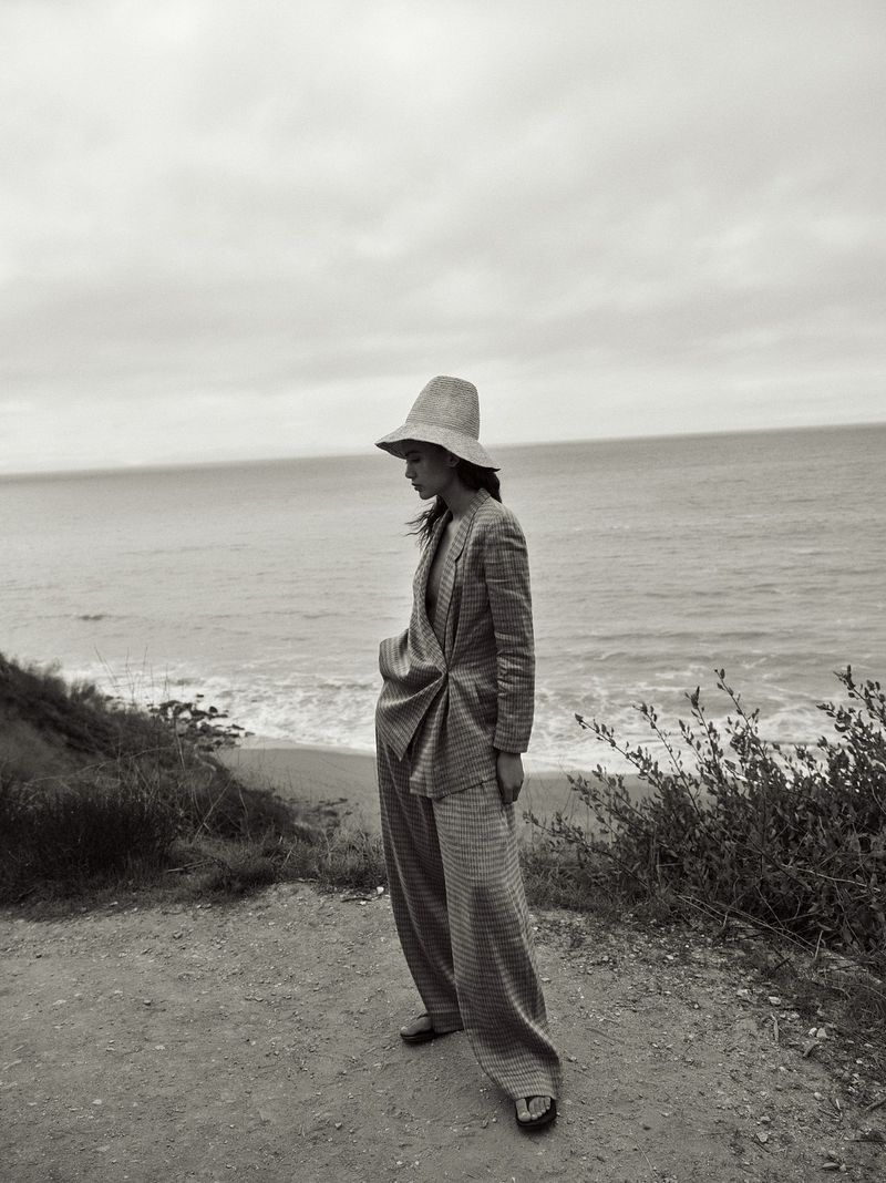 A woman in a pantsuit and hat stands on a cliff overlooking the ocean. The image is black and white, emphasizing the textures of the clothing.