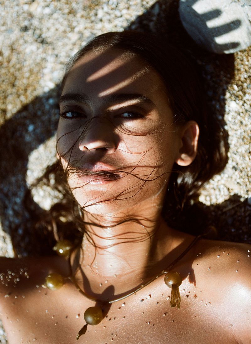 Close-up portrait of a woman with wet hair lying on sand, wearing a necklace, with shadows on her face and warm sunlight on her skin.