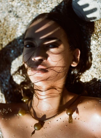 Close-up portrait of a woman with wet hair lying on sand, wearing a necklace, with shadows on her face and warm sunlight on her skin.