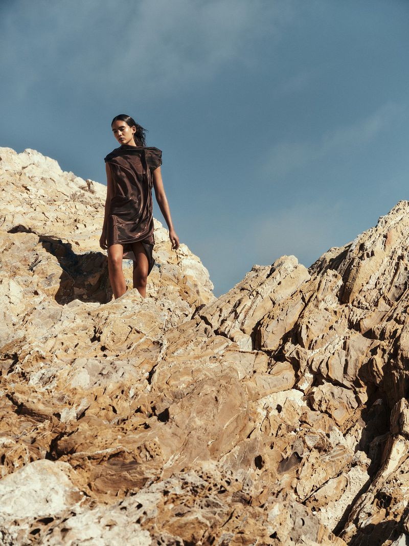 A woman in a brown dress stands on a rocky cliff against a blue sky. A fashion shot featuring outdoor apparel on a dry, rocky landscape.