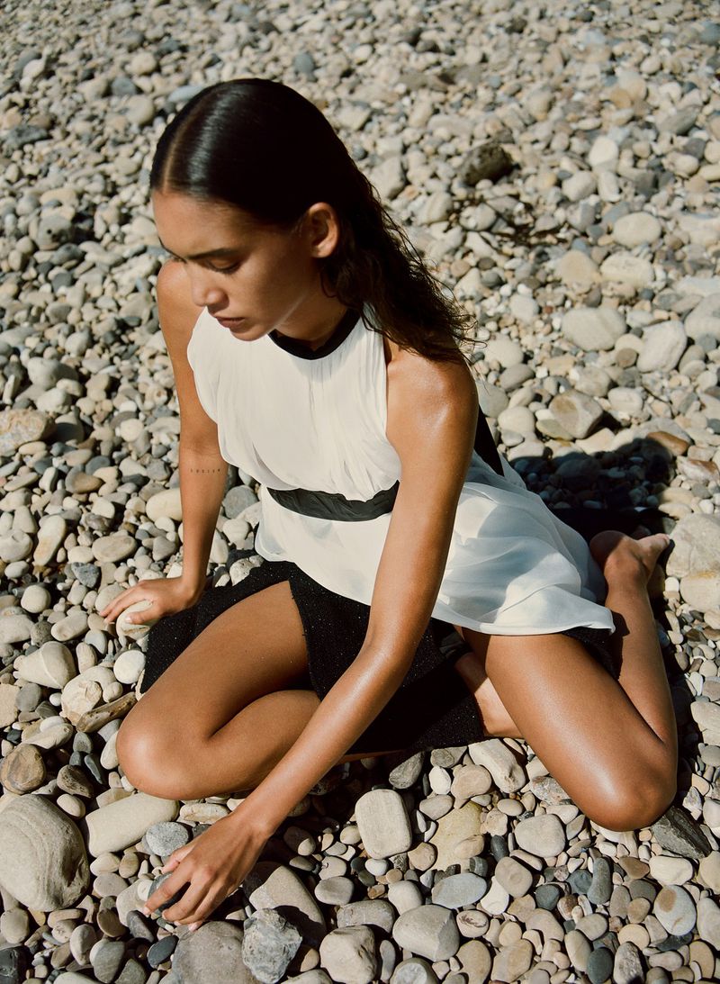 Woman in flowing white dress sits on pebble-covered beach in outdoor fashion shot, gazing down in natural light on sunny day.