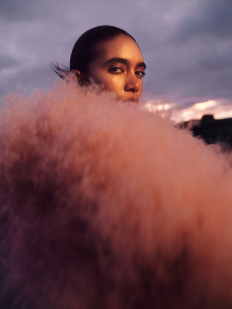 Portrait of an adult female with dark hair partially obscured by a pink smoke effect outdoors with a sunset and cloudy sky.