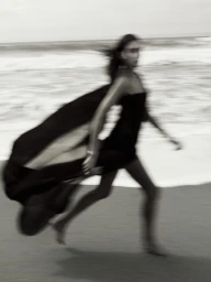 Motion-blurred model in a dark strapless dress running along a sandy beach near ocean waves.