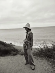Model in wide-brim hat and tailored trousers standing on a coastal cliff path overlooking the ocean