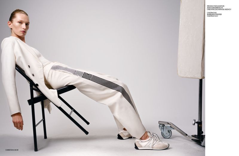 A sleek woman in a stylish white pantsuit and sneakers reclines on a black chair in a bright studio, capturing modern fashion.