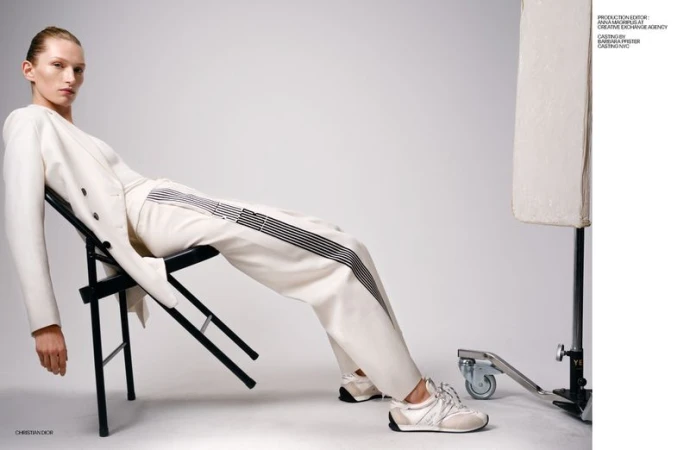 A sleek woman in a stylish white pantsuit and sneakers reclines on a black chair in a bright studio, capturing modern fashion.