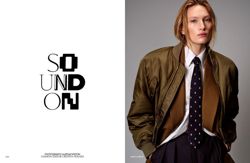 Fashion editorial portrait of an adult woman wearing an olive bomber jacket, white shirt, and navy tie in a photo studio.
