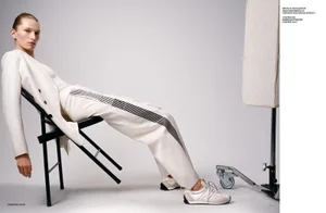 Model Liz Kennedy wearing white attire and sneakers reclining on a black folding chair in a studio