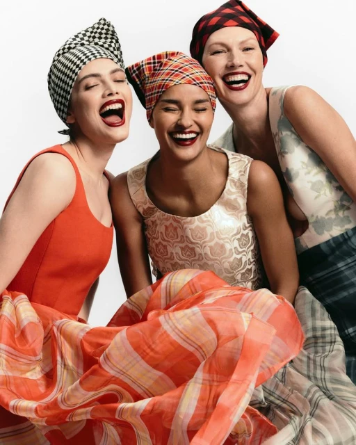 Three adult women laugh together, wearing patterned headscarves and colorful patterned fabrics in a bright, clean indoor setting.
