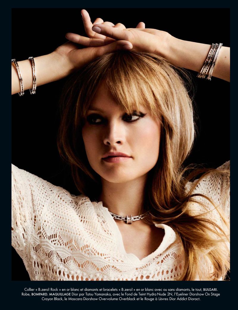 Fashion shot featuring a blond adult female with a white top and silver bracelets raising her hands over her head against a black background.