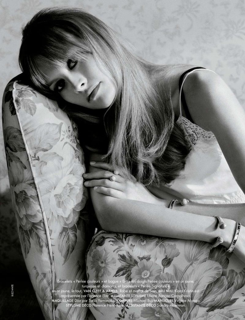 Black and white portrait of a woman with long hair reclining on a floral patterned chair in a studio with natural lighting.