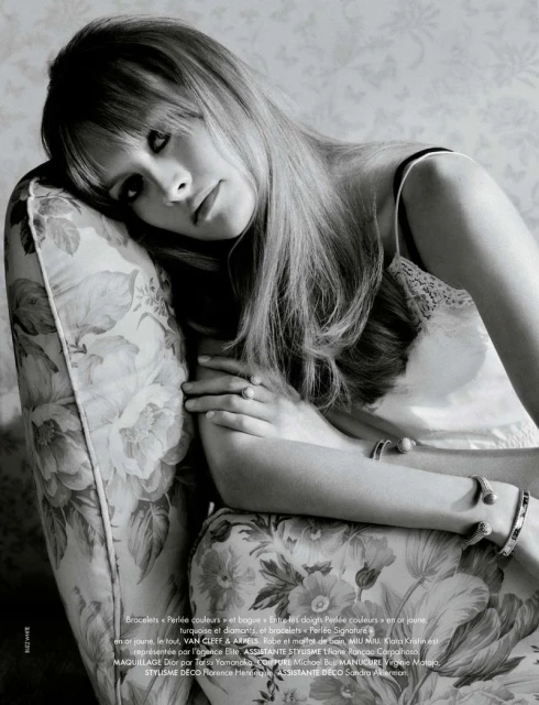 Black and white portrait of a woman with long hair reclining on a floral patterned chair in a studio with natural lighting.