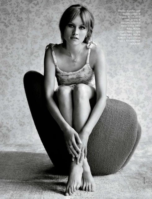 A young woman with blonde hair sits on a gray chair with her legs crossed, set against a wallpapered background in a studio shot.