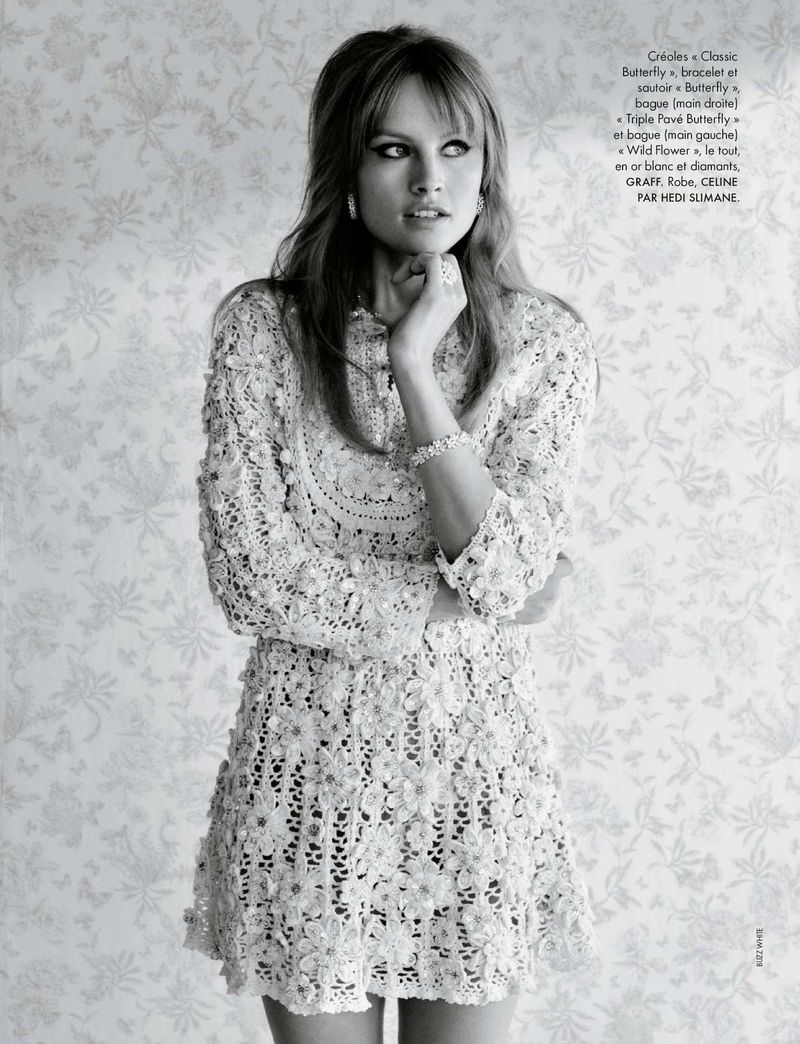 A woman with blonde hair in a white lace dress poses in front of a light floral wallpaper for a fashion or beauty editorial shot.