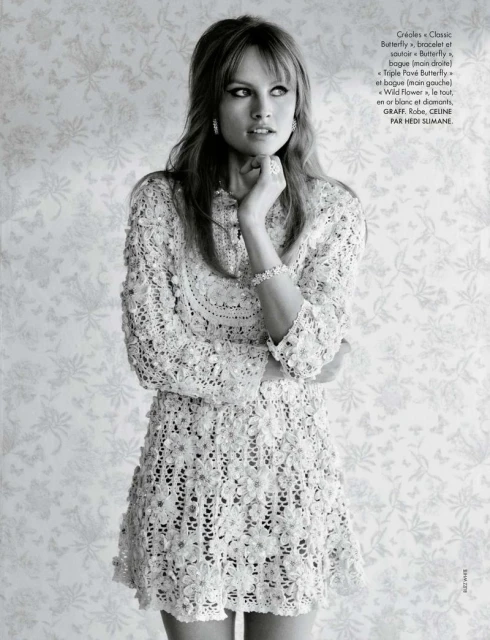 A woman with blonde hair in a white lace dress poses in front of a light floral wallpaper for a fashion or beauty editorial shot.