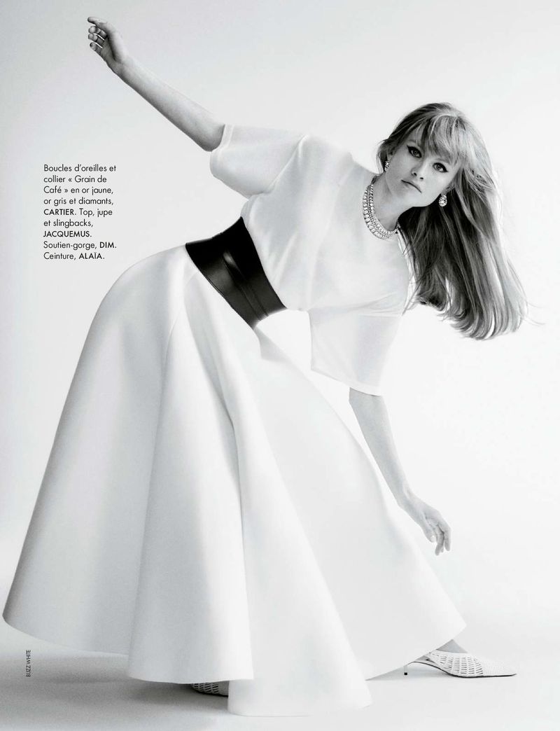 Woman poses in a white dress with a wide black belt, jewelry, and white heels against a bright white studio background for a fashion editorial.
