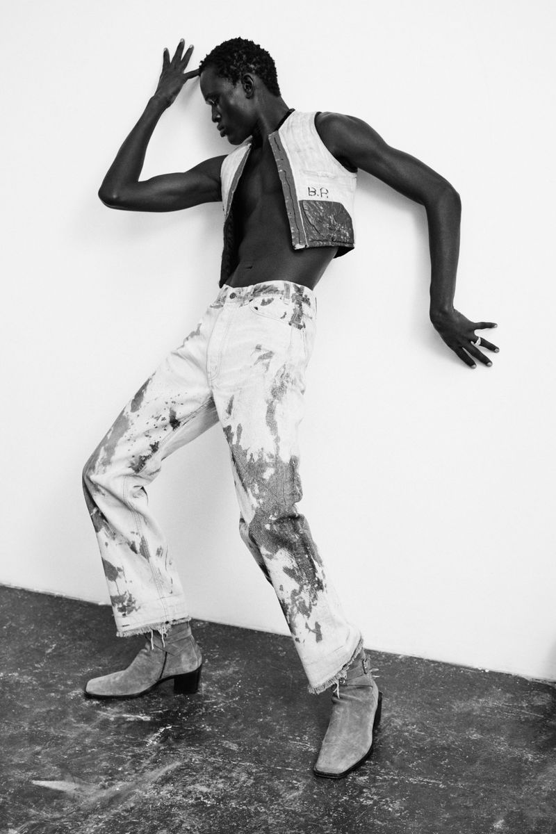 Black and white fashion portrait of a Black male fashion model in a vest and paint-splattered jeans leaning against a wall in boots.