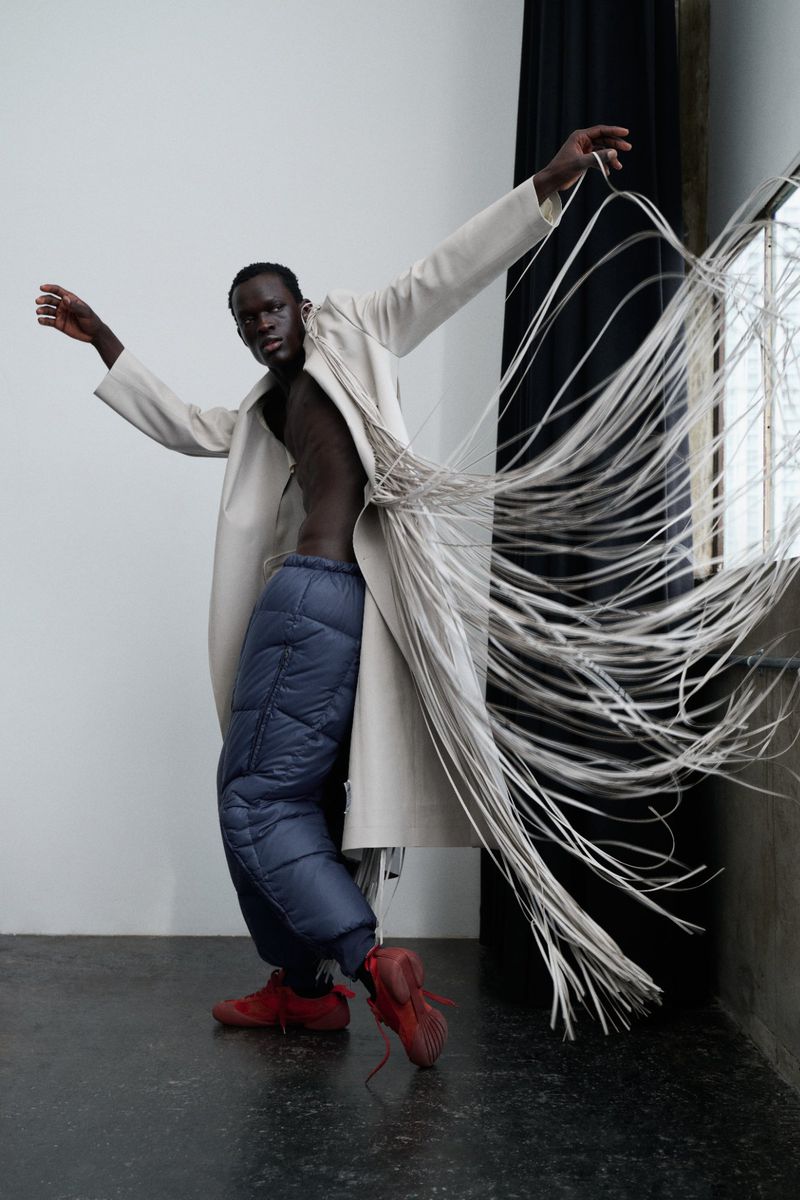 Editorial fashion shot of a Black male model in an open beige coat with fringe, blue quilted pants, and red shoes striking a pose indoors.