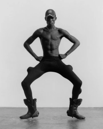 Monochrome portrait of an androgynous person posing in a cap, tights with bulbous extensions, and large boots in a studio setting.