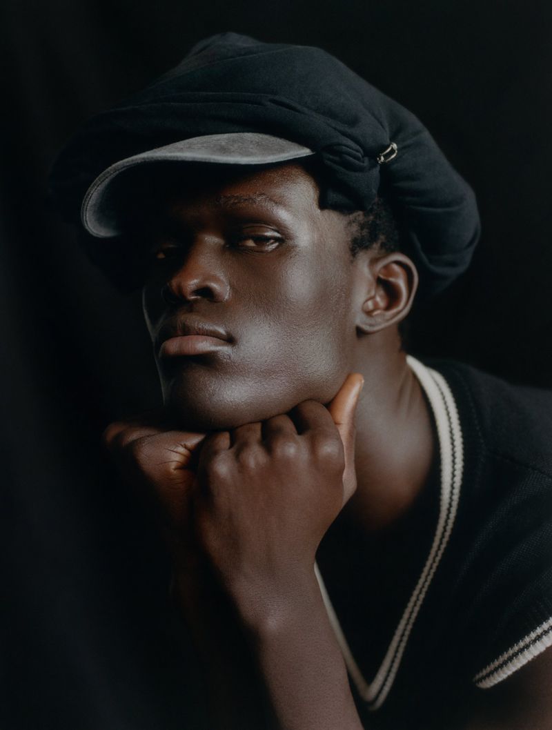 Close-up portrait of a Black male model in a cap, resting his face on his hands, set against a dark background in a photo studio.