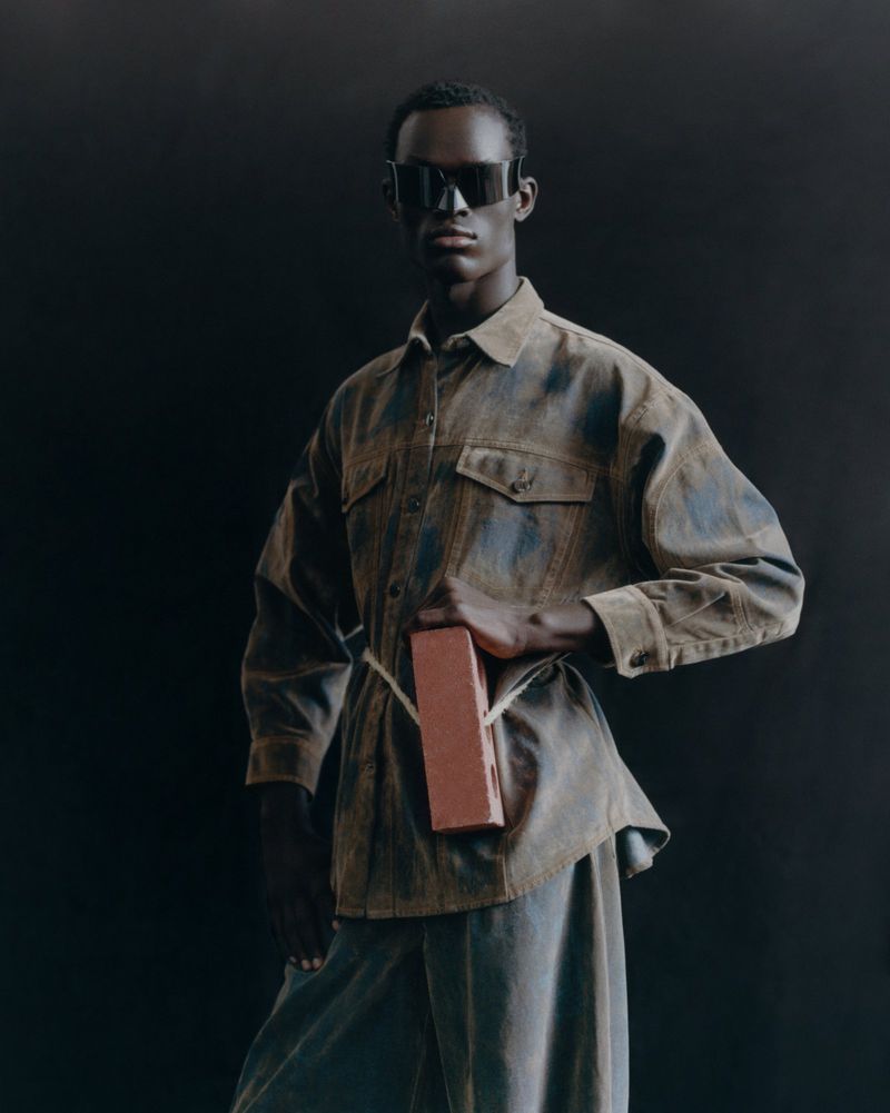 Fashion shot of a dark-skinned man wearing sunglasses and a patterned shirt, holding a brick against a dark background indoors.