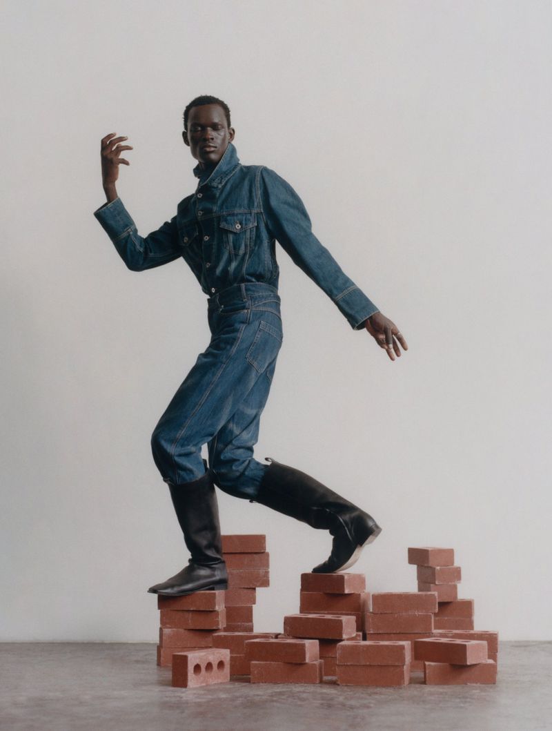A black male fashion model wearing a blue denim outfit and black boots poses atop a small brick staircase in a studio setting.