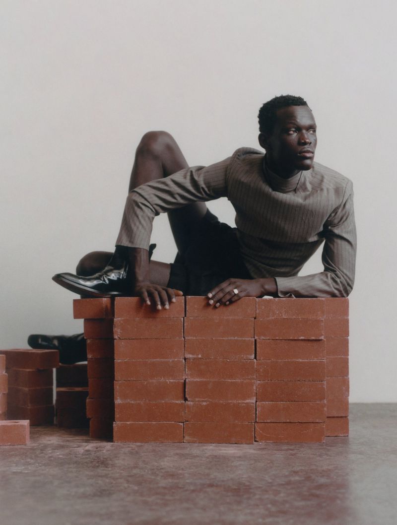Elegant dark-skinned man posing on bricks in a photo studio, wearing a gray suit and shiny black boots, gazing off camera with a neutral expression.