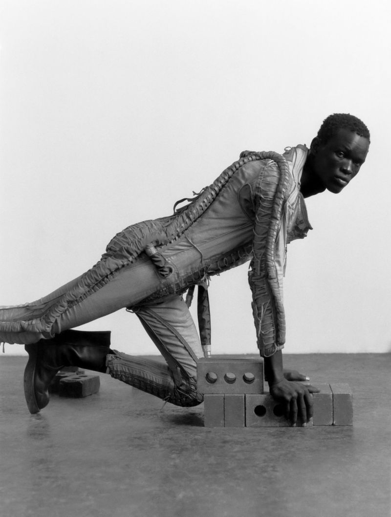 Monochrome fashion shot of a black male model in structured jumpsuit, kneeling on red bricks in a studio setting with a blank backdrop.