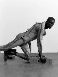 Black model posing on hands and knees over stacked bricks against a plain studio backdrop, wearing textured avant-garde apparel.