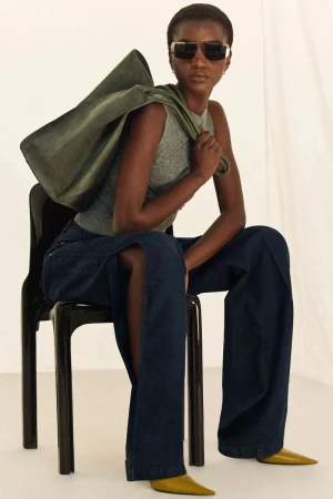 A dark-skinned woman is sitting on a black chair, wearing a sleeveless gray top, wide blue jeans, olive shoes, carrying a matching bag.