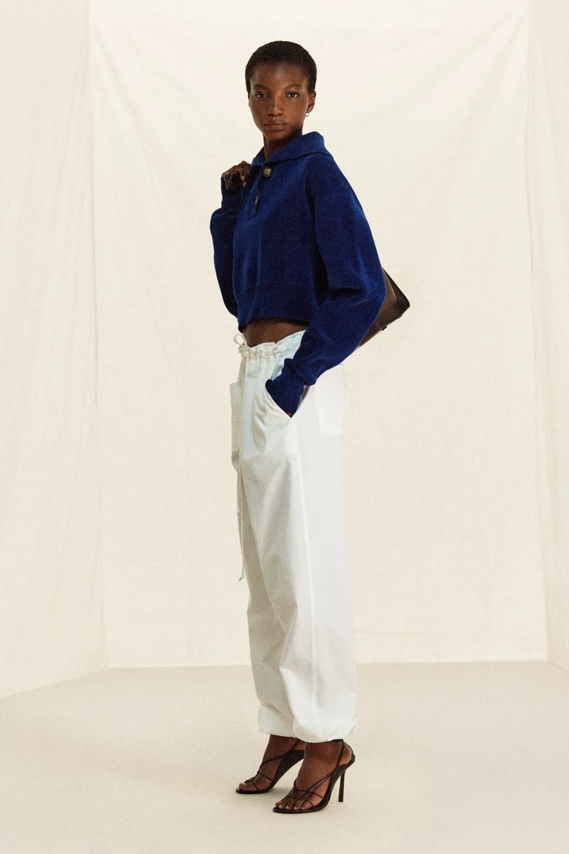 Dark-skinned woman in a blue collared sweater and white cargo pants poses indoors against a neutral backdrop, holding a brown purse.